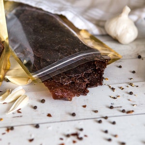 May include: Two bags of dried meat, one gold and one clear, with the clear bag open and the meat spilling out. The meat is dark brown and has a rough texture. There are also some cloves of garlic and black peppercorns scattered on a white wooden surface.