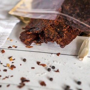May include: A close-up of a bag of beef jerky with a white background. The jerky is dark brown and has a spicy, peppery flavor. The bag is open and the jerky is spilling out. There are also some peppercorns and garlic cloves on the table.