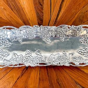 May include: A silver serving tray with a grapevine design. The tray is rectangular and has a raised edge. The grapevine design is intricate and detailed, with leaves and grapes.