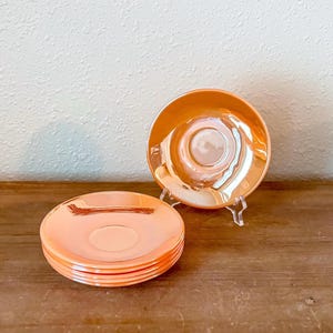 May include: A stack of four orange-colored saucers and one additional saucer displayed on a clear stand. The saucers have a glossy, iridescent finish, reflecting light with a warm, peachy hue. The saucers are likely for a tea or coffee set.
