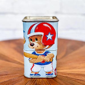 May include: A vintage tin piggy bank featuring a cartoon bear wearing a baseball uniform and a helmet. The bear is holding a baseball bat and is depicted on a blue background with white accents.