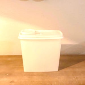 May include: A white, rectangular food storage container with a matching lid. The lid has a small, round opening. The container is made of a solid, opaque material and sits on a wooden surface.