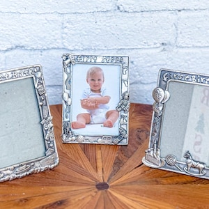 May include: Three silver-toned picture frames with a birthday theme. The frames have a raised design with various birthday-related images, including balloons, presents, and a cake. The frames are all empty and ready for photos.