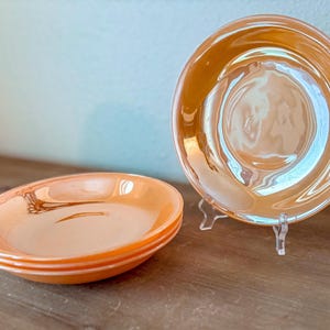 May include: A set of three iridescent orange glass plates, with a fourth plate displayed on a clear stand. The plates have a glossy finish and a subtle, reflective sheen. The plates are on a wooden surface.