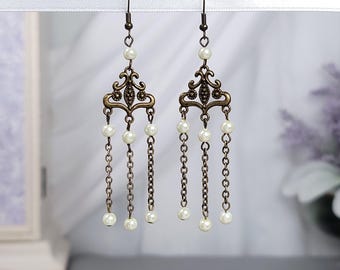 May include: Ornate chandelier earrings with a bronze finish. Each earring features a decorative filigree top, dangling chains, and faux pearl accents. The earrings are designed to be worn with pierced ears.