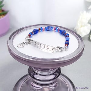 May include: A silver bracelet with the word "DIABETIC" engraved on a rectangular plate. The bracelet features blue and red beads. The bracelet is displayed on a purple pedestal.