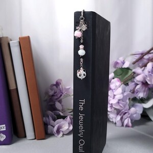 May include: A silver bookmark with a panda charm, pink pom-pom, and white bead accents. The bookmark is attached to a black book with the text "The Jewelry Owl". The background includes other books and purple flowers.