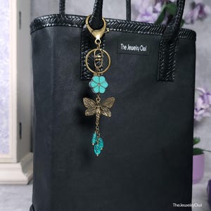 May include: A black tote bag with a decorative keychain. The keychain features a turquoise flower, a bronze dragonfly, and dangling blue beads. The keychain is attached to a gold-colored clasp. The text "The Jewelry Owl" is visible on the bag.
