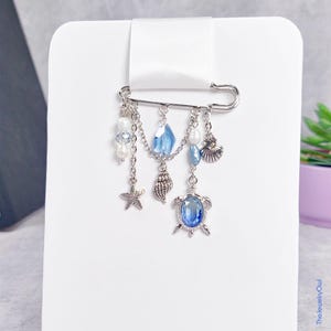 May include: A silver-toned safety pin brooch featuring dangling charms including a blue crystal turtle, a starfish, a seashell, and white pearls. The brooch is attached to a white ribbon on a white display card. The text TheJewelryOwl is visible.