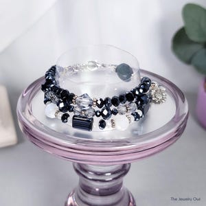 May include: A stack of beaded bracelets in shades of black, clear, and white, with silver accents. The bracelets are displayed on a small, light purple pedestal. One bracelet has a silver charm. The Jewelry Owl logo is in the bottom right corner.