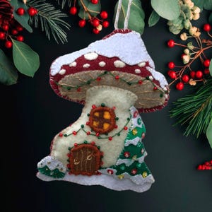 Op de afbeelding: Een vilten kerstversiering in de vorm van een paddenstoel. De bordeauxrode paddenstoelhoed heeft witte stippen en een besneeuwde rand. De witte steel heeft een bruine deur en een raam. Een kleine groene boom en een slinger van rode en groene kralen versieren de versiering.