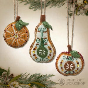 May include: Three handmade felt fruit ornaments: an orange slice, a pear, and an apple. Each is decorated with beads and embroidery, with a brown stem and green felt leaves. They are suspended from twine. The ornaments are festive.