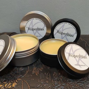 Może przedstawiać: Cztery małe metalowe puszki z woskiem pszczelim, każda z białą etykietą z napisem "RopesWax". Puszki znajdują się na brązowej skórzanej powierzchni.