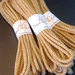 Może przedstawiać: Trzy wiązki liny w naturalnym kolorze, ciasno zwinięte i zabezpieczone białymi etykietami. Etykiety zawierają tekst "Ougon" i "Andrea Ropes" w okrągłym wzorze. Lina wydaje się być wykonana z grubego, włóknistego materiału.