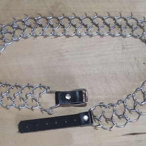 Op de afbeelding: Een zilveren kettinghondensnoer met scherpe punten en een zwarte leren riem met een gesp.