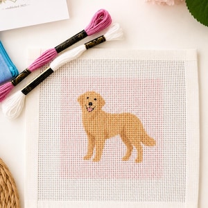 May include: A square needlepoint canvas featuring a golden retriever dog design in shades of gold, black, and pink, accompanied by two skeins of embroidery floss in magenta and white, with a small card showing a white dog illustration.