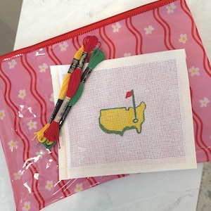 Puede incluir: Un kit de punto de aguja con un dise&ntilde;o de mapa amarillo y verde con una bandera roja, junto con hilo de bordar amarillo, rojo y verde. El kit est&aacute; colocado sobre una bolsa con cremallera rosa con estampado floral.