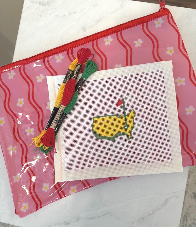 Puede incluir: Un kit de punto de aguja con un dise&ntilde;o de mapa amarillo y verde con una bandera roja, junto con hilo de bordar amarillo, rojo y verde. El kit est&aacute; colocado sobre una bolsa con cremallera rosa con estampado floral.