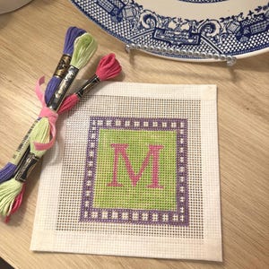 May include: A needlepoint canvas with a green center and a pink letter "M" is displayed. The canvas is surrounded by a purple border with a dotted pattern. Skeins of embroidery floss in purple, green, and pink are tied together with a pink ribbon.