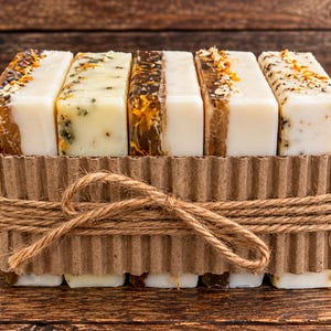 May include: Five rectangular bars of soap arranged in a row inside a corrugated cardboard sleeve tied with twine. Each bar features a dual-layered design with one side being opaque white and the other translucent amber, topped with dried botanical inclusions.