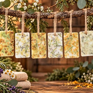 May include: Six rectangular wax tablets hang from a rustic wooden branch by jute twine. Each white wax bar is embedded with various dried botanical elements, including green herbs, yellow flower petals, and small purple lavender sprigs.