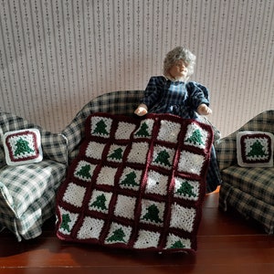 Puede incluir: Una escena en miniatura con una muñeca sentada en un sofá a cuadros, flanqueada por sillones. Se ve una manta de ganchillo con árboles de Navidad verdes sobre cuadrados blancos y una almohada a juego. La escena está ambientada sobre un fondo de papel pintado.