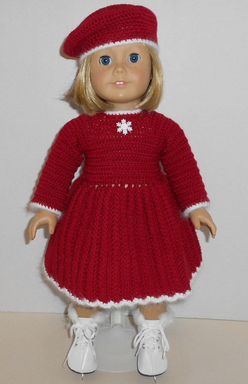 Puede incluir: Un vestido rojo de ganchillo con un dise&ntilde;o de copo de nieve blanco y ribete blanco. El vestido lo lleva una mu&ntilde;eca con el pelo rubio y los ojos azules. La mu&ntilde;eca lleva una boina roja de ganchillo con ribete blanco. La mu&ntilde;eca tambi&eacute;n lleva patines de hielo blancos.