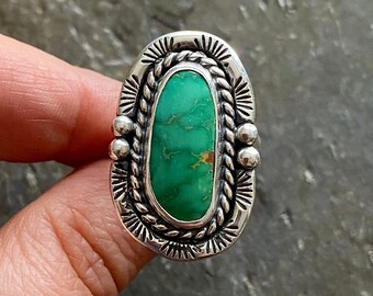 May include: A silver ring with a green turquoise stone set in a detailed silver bezel. The ring has a rope-like design around the stone and small silver beads on the sides.
