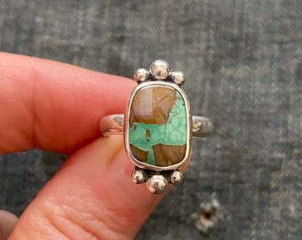 May include: A silver ring featuring a rectangular stone with a matrix of turquoise and brown patterns. The stone is set in a smooth silver bezel with three small silver spheres at the top and bottom. A plain silver band completes the ring design.