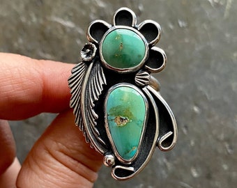 May include: A silver ring with two turquoise stones set in a floral design. The ring is on a finger.