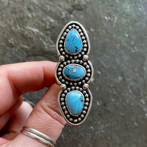 May include: A silver ring featuring three turquoise stones. The ring has a vertical design with a beaded border around each stone. The turquoise stones are oval and teardrop-shaped, with a light blue color and dark blue veining.