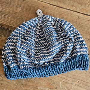 May include: A knitted beanie hat in shades of blue and white, with a textured zigzag pattern. The hat has a solid blue brim and a small gray loop at the top. The hat is made of soft yarn.