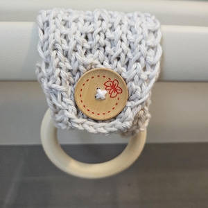May include: A beige knitted loop with a wooden button featuring a red flower design. The loop is attached to a white wooden ring.