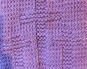 May include: Close-up of a hand-knitted lavender-colored square with a textured pattern. The knitted fabric features a central design, possibly a cross or a stylized shape, within the overall knit. The texture is visible, with each stitch clearly defined.