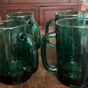 May include: Four emerald green glass mugs with handles. The mugs have a rectangular shape with rounded corners and a gold rim. The mugs are arranged on a wooden surface.
