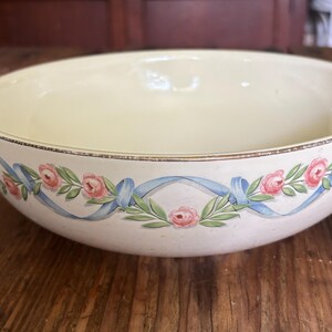 May include: Cream-colored ceramic bowl with a floral design. The bowl features a decorative border of pink roses, blue ribbons, and green leaves. The rim has a thin gold trim. The bowl is sitting on a wooden surface.