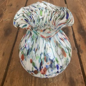 May include: A decorative glass vase with a white base and a ruffled rim. The vase is adorned with colorful flecks of red, blue, green, and orange. The vase has a round body and a wide, flared opening.