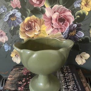 May include: A sage green, pedestal-style bowl with a scalloped rim. The bowl is set against a backdrop of a floral painting with pink, yellow, and blue flowers. The book "Container Gardening Through the Year" is visible.
