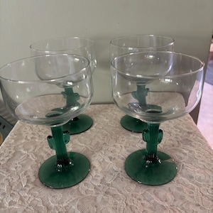May include: Four clear glass margarita glasses with green cactus-shaped stems and bases. The glasses have a wide, rounded bowl. The green stems and bases add a decorative touch to the glassware.