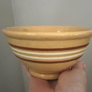 May include: A tan ceramic mixing bowl with a rounded base and a wide rim. The bowl features three horizontal stripes in alternating colors of brown and white. The bowl is held by a hand.