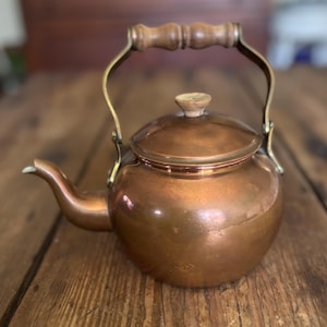 May include: A polished copper tea kettle with a rounded body, a curved spout, and a removable lid featuring a small wooden knob. The kettle includes a brass-toned metal handle with a contoured wooden grip attached to the sides of the vessel.