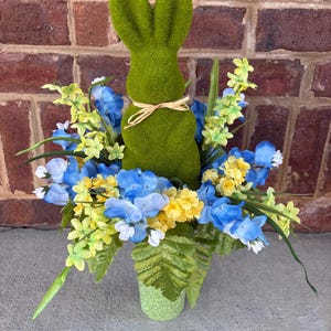 May include: A green, moss-covered bunny figurine with a raffia bow, surrounded by blue, yellow, and white artificial flowers and greenery. The arrangement is in a green, speckled vase, perfect for Easter decor.