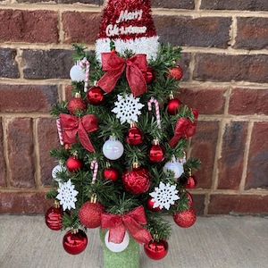 May include: A small artificial Christmas tree decorated with red and white ornaments, including shiny red spheres, glittery red bows, white snowflake cutouts, and miniature candy canes. A red tinsel Santa hat with Merry Christmas text sits on top.