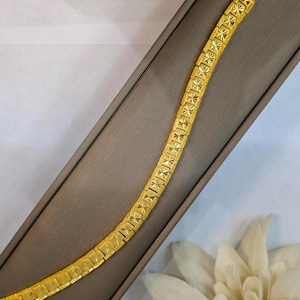 Könnte beinhalten: Ein goldfarbenes Armband mit einem sich wiederholenden Sternmuster. Das Armband ist rechteckig und hat einen Hakenverschluss. Es wird auf einer braunen Oberfläche mit einer weißen Blume im Hintergrund präsentiert.