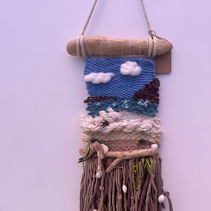 May include: A woven wall hanging with a blue sky, white clouds, and brown mountains. The hanging has a brown fringe with white beads and driftwood accents.