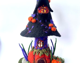 Könnte beinhalten: Eine skurrile Halloween-Dekoration in Form eines Hexenhut-Hauses. Der schwarze Hut ist mit orangefarbenen Kürbissen und lila Ranken verziert. Das Haus hat einen roten und violetten Sockel mit einer kleinen Tür und einem Fenster.