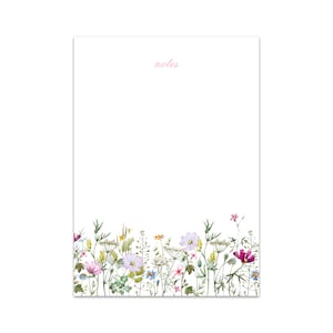 Puede incluir: Un bloc de notas blanco con la palabra "notes" impresa en rosa en la parte superior. La parte inferior del bloc de notas presenta un borde de flores silvestres de acuarela en tonos de rosa, morado, amarillo y verde.