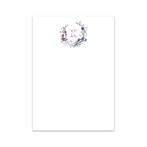 Puede incluir: Un bloc de notas blanco con un diseño de corona floral con flores rosas, azules y moradas. La letra "K" está en el centro de la corona en un color rosa claro.