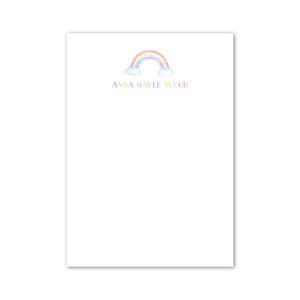 Puede incluir: Un bloc de notas blanco con un diseño de arcoíris pastel y el texto "ANNA GAYLE WOOD" en una fuente pastel a juego. El arcoíris está arqueado sobre dos nubes blancas y esponjosas. El bloc de notas es rectangular.