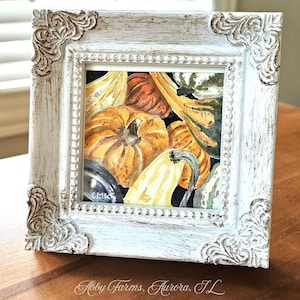 May include: A square watercolor painting of various orange and green pumpkins and gourds set within a distressed white wooden frame with ornate corner detailing. The painting features the initials CMK in the bottom left corner.